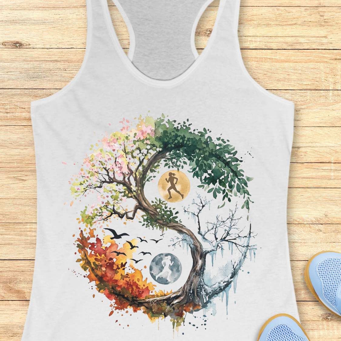 A white racerback tank top laid flat on a light wood plank background, featuring a large colorful watercolor-style graphic of a yin-yang tree with autumn leaves, birds, moon phases, and a running figure. Part of a blue shoe is visible at the bottom right.
