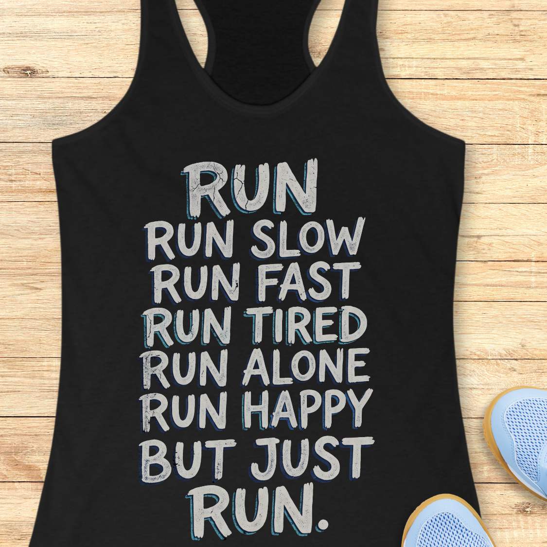A black racerback tank top with a motivational running quote printed in white and light blue text, laid flat on a light wood background with blue running shoes partially visible at the bottom right.