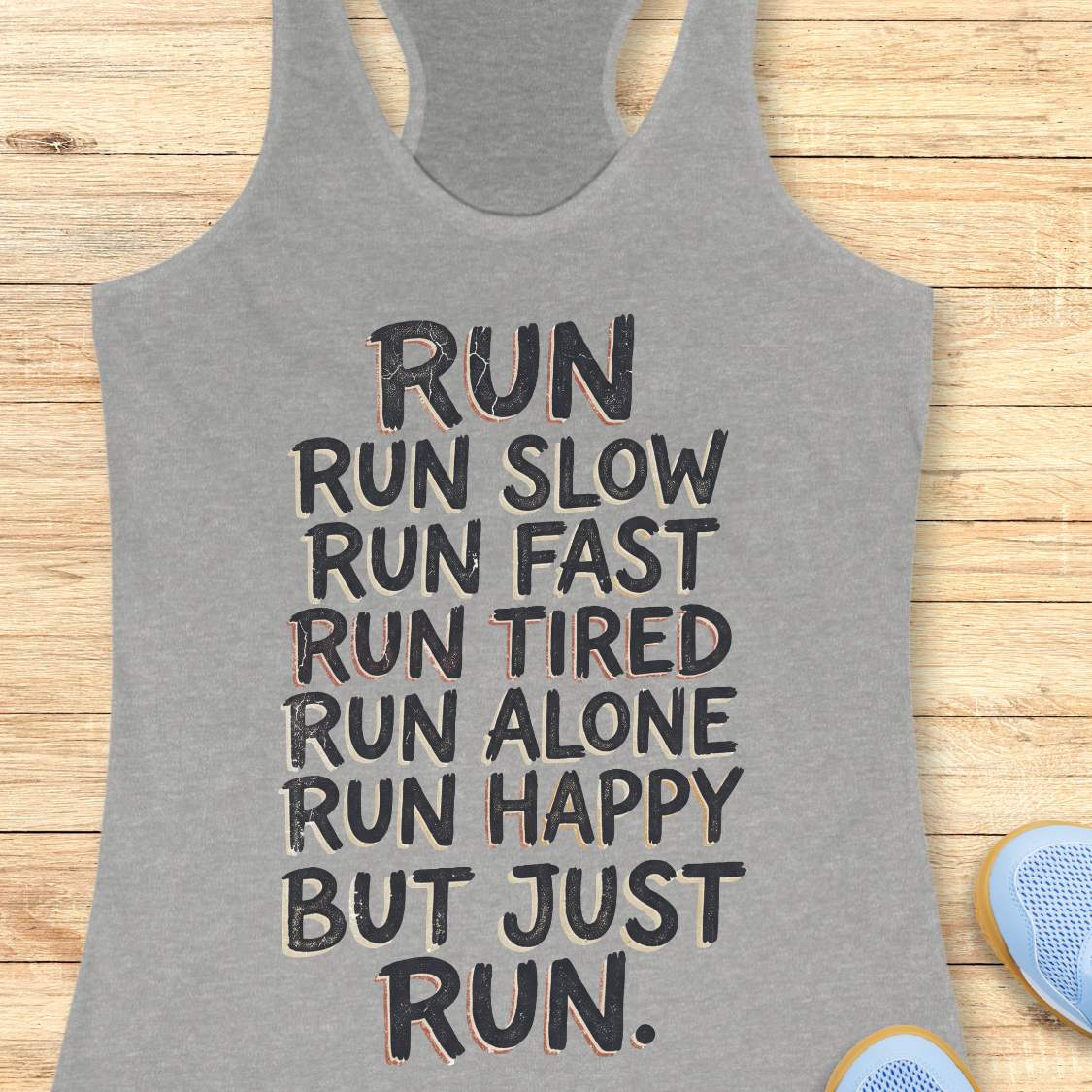 A heather gray racerback tank top with a motivational quote printed on the front, laid flat on a light wood background with partial blue sneakers visible at the bottom right.