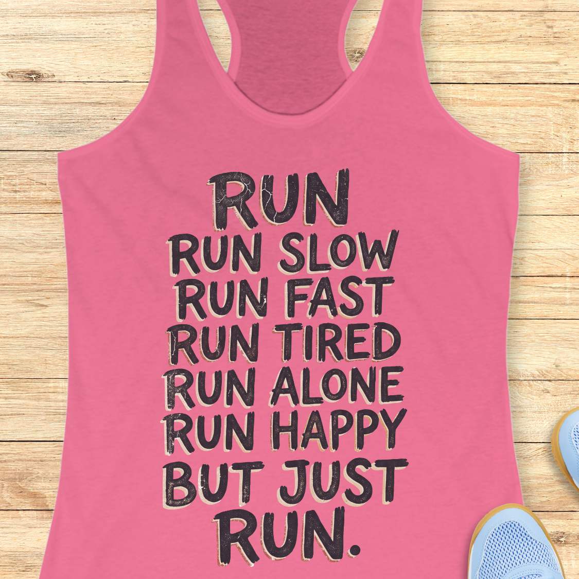 A pink racerback tank top with black motivational text reading 'RUN RUN SLOW RUN FAST RUN TIRED RUN ALONE RUN HAPPY BUT JUST RUN.' laid flat on a light wood background, with partial blue shoes visible at the edges.