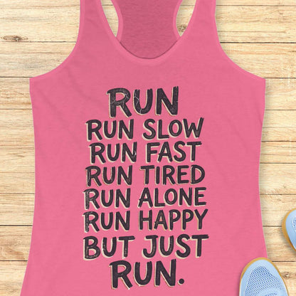 A pink racerback tank top with black motivational text reading 'RUN RUN SLOW RUN FAST RUN TIRED RUN ALONE RUN HAPPY BUT JUST RUN.' laid flat on a light wood background, with partial blue shoes visible at the edges.