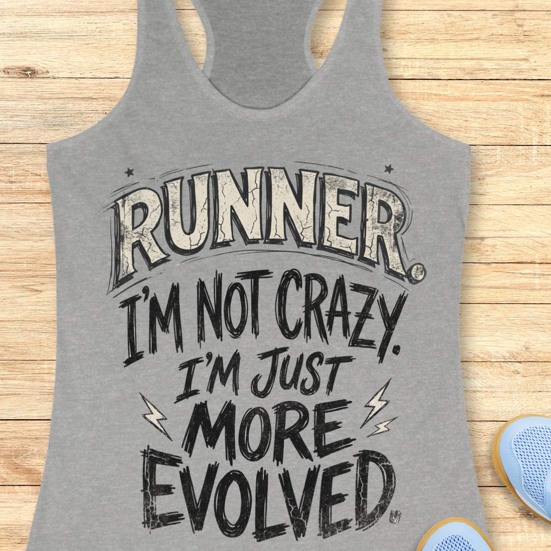 A heather gray racerback tank top with a humorous graphic print reading 'RUNNER I'M NOT CRAZY I'M JUST MORE EVOLVED' in distressed black and white lettering, laid flat on a light wood background with partial blue sneakers visible at the bottom right.