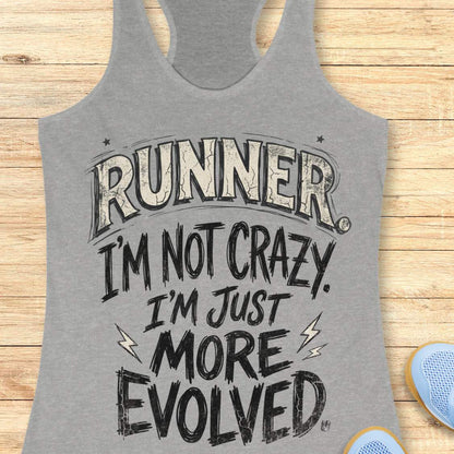 A heather gray racerback tank top with a humorous graphic print reading 'RUNNER I'M NOT CRAZY I'M JUST MORE EVOLVED' in distressed black and white lettering, laid flat on a light wood background with partial blue sneakers visible at the bottom right.
