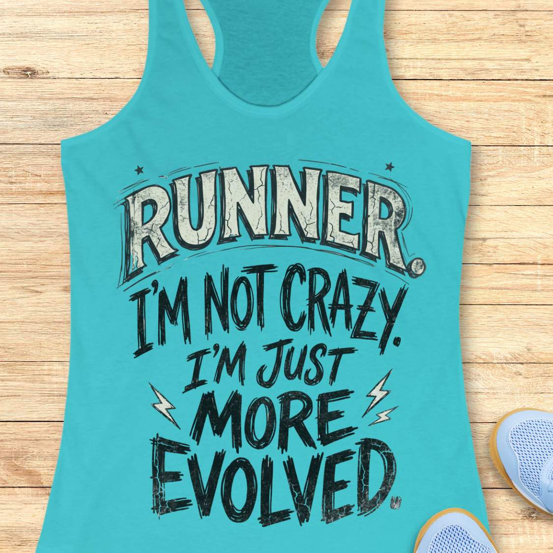 A turquoise racerback tank top laid flat on a light wood surface, printed with the phrase “RUNNER I’M NOT CRAZY I’M JUST MORE EVOLVED” in distressed white and black lettering. Partial blue sneakers are visible at the lower right as styling props.