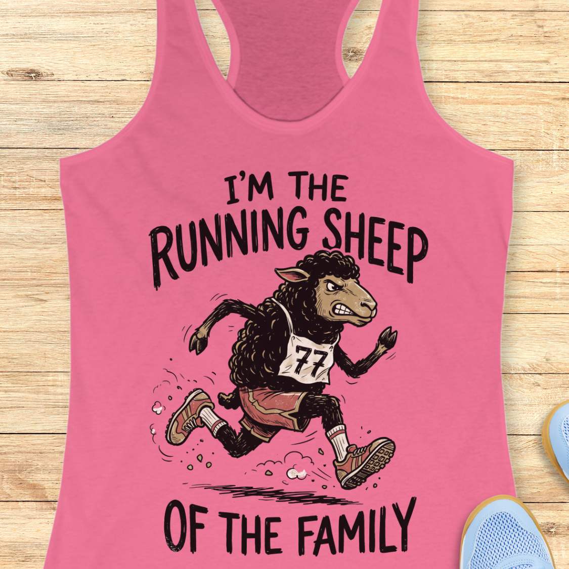 A pink racerback tank top laid flat on a light wood surface, featuring a cartoon running sheep graphic with the text 'I'M THE RUNNING SHEEP OF THE FAMILY.' A small blue shoe is partially visible at the bottom right as a styling prop.