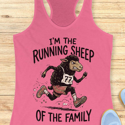 A pink racerback tank top laid flat on a light wood surface, featuring a cartoon running sheep graphic with the text 'I'M THE RUNNING SHEEP OF THE FAMILY.' A small blue shoe is partially visible at the bottom right as a styling prop.