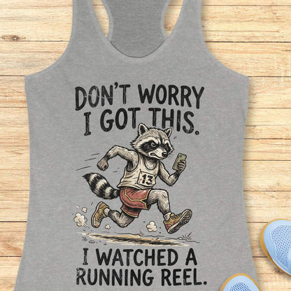 A heather gray racerback tank top laid flat on a light wood surface, featuring a humorous graphic of a raccoon running in a race with the text 'DON'T WORRY I GOT THIS. I WATCHED A RUNNING REEL.
