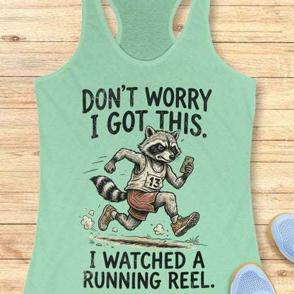 A mint green racerback tank top laid flat on a light wood background, featuring a humorous graphic of a raccoon running a race with the text 'DON'T WORRY I GOT THIS. I WATCHED A RUNNING REEL.