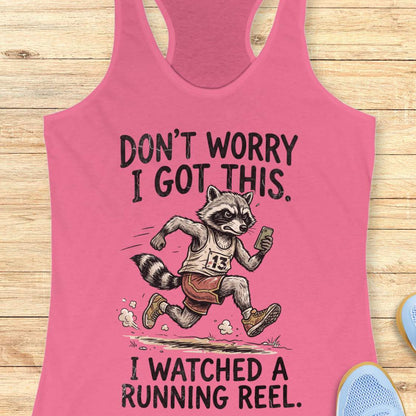 A pink racerback tank top laid flat on a light wood background, featuring a humorous graphic of a raccoon running while holding a phone, with the text 'DON'T WORRY I GOT THIS.' above and 'I WATCHED A RUNNING REEL.