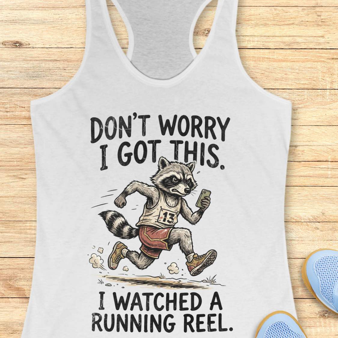 A white racerback tank top laid flat on a light wood background, featuring a humorous graphic of a raccoon running with the text 'DON'T WORRY I GOT THIS. I WATCHED A RUNNING REEL.
