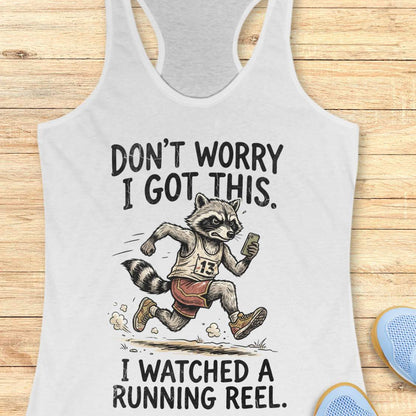 A white racerback tank top laid flat on a light wood background, featuring a humorous graphic of a raccoon running with the text 'DON'T WORRY I GOT THIS. I WATCHED A RUNNING REEL.