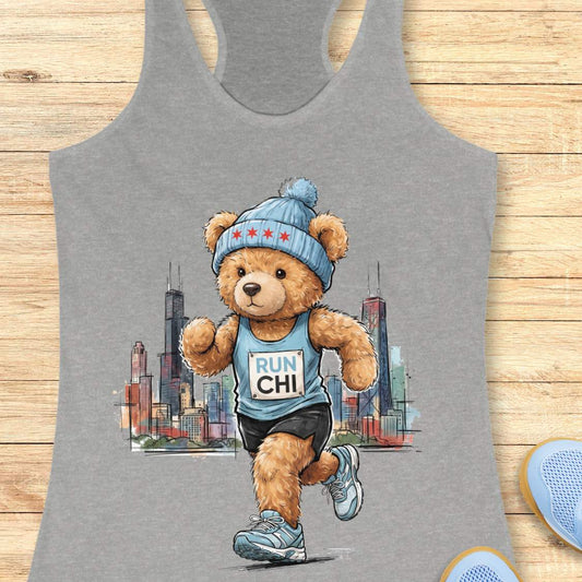 Heather gray racerback running tank top featuring a watercolor-style illustration of a teddy bear running in front of the Chicago skyline. The bear wears a blue knit beanie decorated with red stars, a blue running singlet with a race bib that reads “RUN CHI,” black running shorts, and blue running shoes. Behind the bear is a sketch-style cityscape inspired by Chicago, including prominent skyscrapers. The tank top is displayed on a light wood background with partial running shoes visible in the corners.