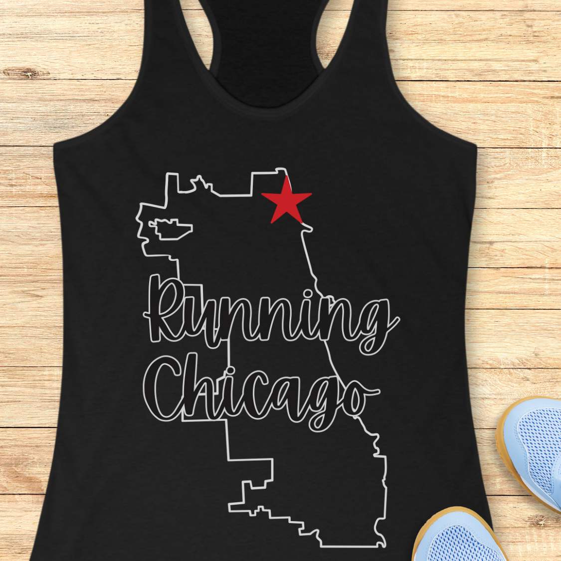 Black racerback running tank top featuring a minimalist white outline of the city of Chicago. A large red six-pointed star, inspired by the Chicago flag, marks a location near the upper right section of the city outline. The words “Running Chicago” appear in large script lettering layered over the map design. The tank top is displayed on a light wood background with partial blue running shoes visible in the bottom corners. The design has a clean, modern look celebrating running in Chicago.