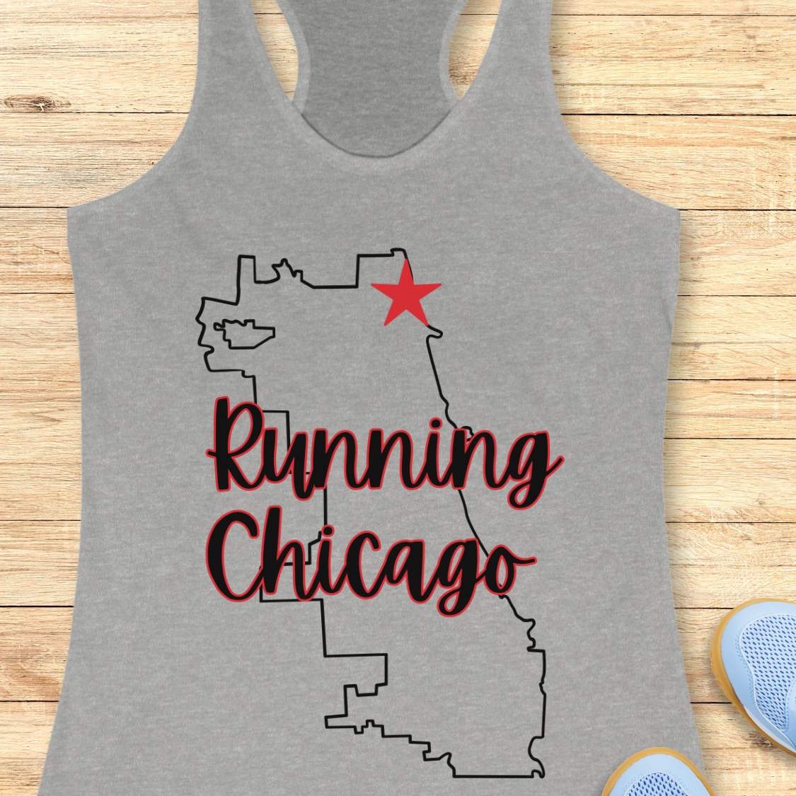 grey racerback running tank top featuring a minimalist white outline of the city of Chicago. A large red six-pointed star, inspired by the Chicago flag, marks a location near the upper right section of the city outline. The words “Running Chicago” appear in large script lettering layered over the map design. The tank top is displayed on a light wood background with partial blue running shoes visible in the bottom corners. The design has a clean, modern look celebrating running in Chicago.