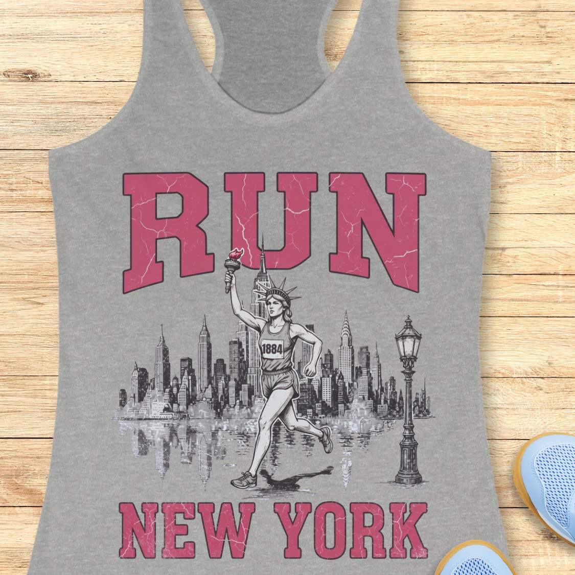 Heather gray racerback running tank top featuring a vintage-style New York running design. Large distressed pink varsity letters spell “RUN” above and “NEW YORK” below a detailed black-and-white illustration of the New York City skyline. In the center, the Statue of Liberty is depicted as a runner, wearing a race bib numbered 1884 and carrying the torch while running in front of the skyline. A classic New York streetlamp appears beside the skyline, which is reflected in water beneath it. The tank top is dis