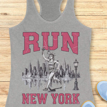Heather gray racerback running tank top featuring a vintage-style New York running design. Large distressed pink varsity letters spell “RUN” above and “NEW YORK” below a detailed black-and-white illustration of the New York City skyline. In the center, the Statue of Liberty is depicted as a runner, wearing a race bib numbered 1884 and carrying the torch while running in front of the skyline. A classic New York streetlamp appears beside the skyline, which is reflected in water beneath it. The tank top is dis