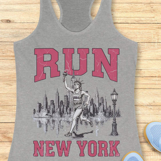 Heather gray racerback running tank top featuring a vintage-style New York running design. Large distressed pink varsity letters spell “RUN” above and “NEW YORK” below a detailed black-and-white illustration of the New York City skyline. In the center, the Statue of Liberty is depicted as a runner, wearing a race bib numbered 1884 and carrying the torch while running in front of the skyline. A classic New York streetlamp appears beside the skyline, which is reflected in water beneath it. The tank top is dis