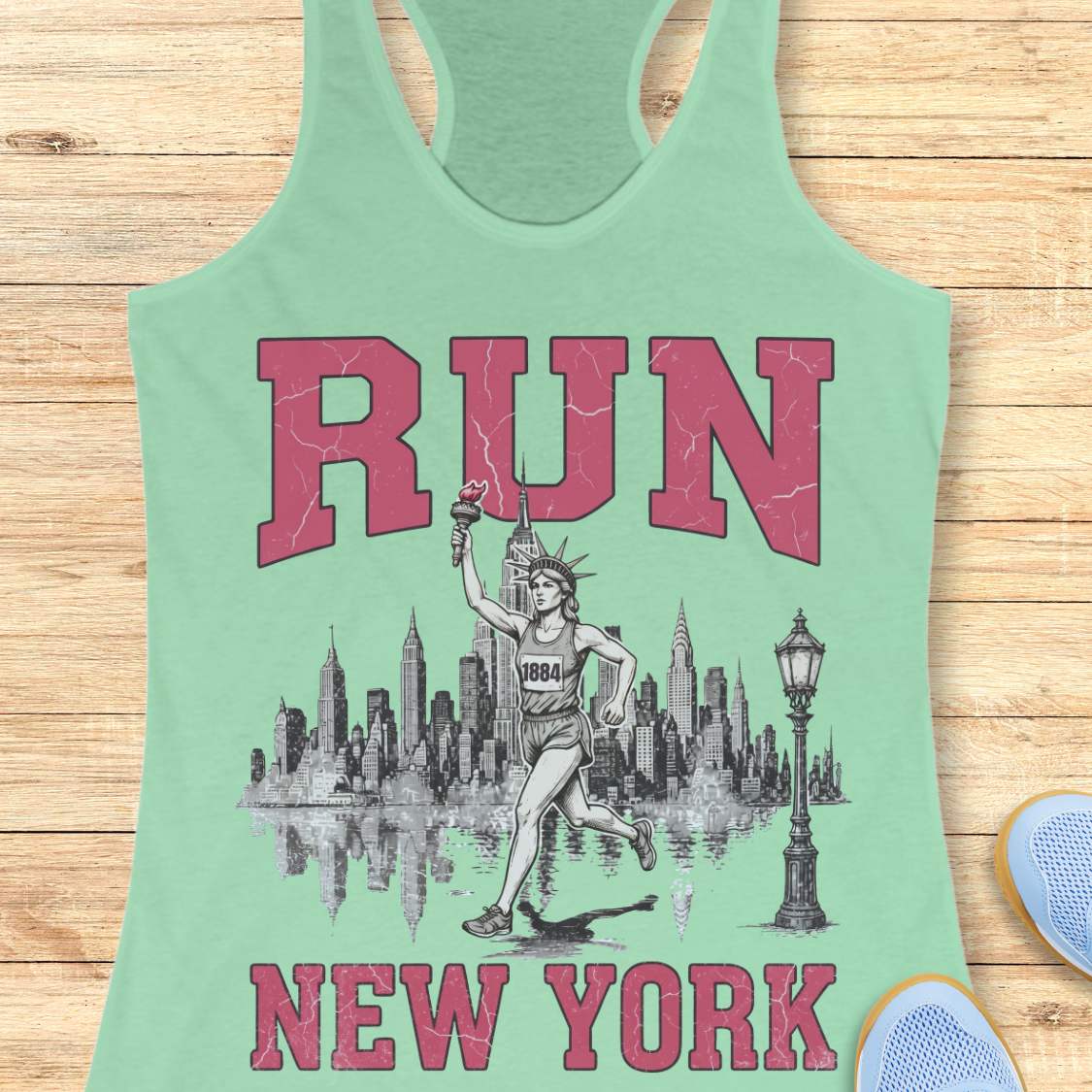 mint racerback running tank top featuring a vintage-style New York running design. Large distressed pink varsity letters spell “RUN” above and “NEW YORK” below a detailed black-and-white illustration of the New York City skyline. In the center, the Statue of Liberty is depicted as a runner, wearing a race bib numbered 1884 and carrying the torch while running in front of the skyline. A classic New York streetlamp appears beside the skyline, which is reflected in water beneath it. The tank top is dis