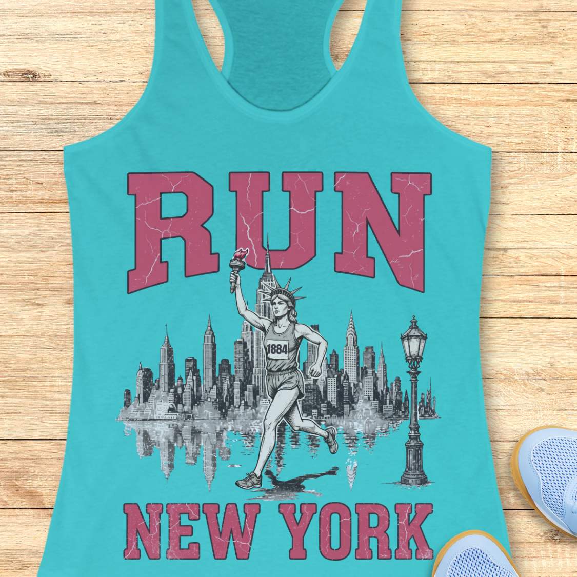 blue racerback running tank top featuring a vintage-style New York running design. Large distressed pink varsity letters spell “RUN” above and “NEW YORK” below a detailed black-and-white illustration of the New York City skyline. In the center, the Statue of Liberty is depicted as a runner, wearing a race bib numbered 1884 and carrying the torch while running in front of the skyline. A classic New York streetlamp appears beside the skyline, which is reflected in water beneath it. The tank top is dis