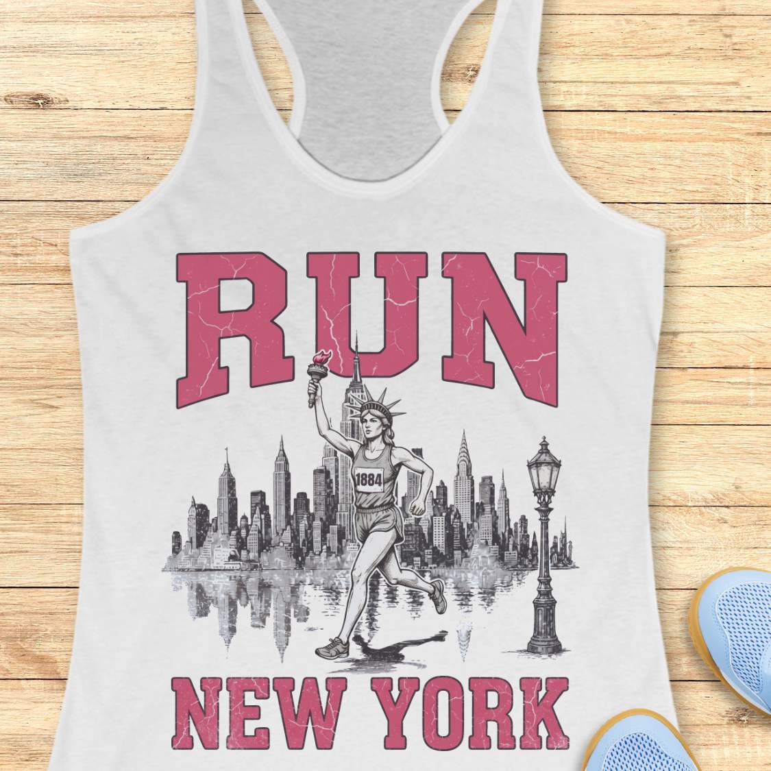white racerback running tank top featuring a vintage-style New York running design. Large distressed pink varsity letters spell “RUN” above and “NEW YORK” below a detailed black-and-white illustration of the New York City skyline. In the center, the Statue of Liberty is depicted as a runner, wearing a race bib numbered 1884 and carrying the torch while running in front of the skyline. A classic New York streetlamp appears beside the skyline, which is reflected in water beneath it. The tank top is dis
