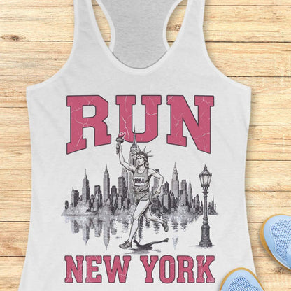 white racerback running tank top featuring a vintage-style New York running design. Large distressed pink varsity letters spell “RUN” above and “NEW YORK” below a detailed black-and-white illustration of the New York City skyline. In the center, the Statue of Liberty is depicted as a runner, wearing a race bib numbered 1884 and carrying the torch while running in front of the skyline. A classic New York streetlamp appears beside the skyline, which is reflected in water beneath it. The tank top is dis