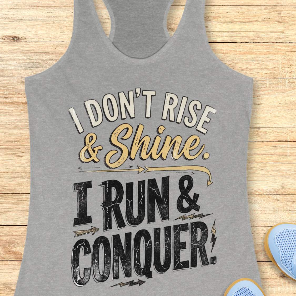 grey racerback running tank top displayed on a light wood background, featuring a distressed vintage-style graphic with blue and white lettering that reads: “I DON’T RISE & SHINE. I RUN & CONQUER.” Decorative arrows and lightning bolt accents surround the text. Partial blue running shoes are visible in the bottom corners of the image.