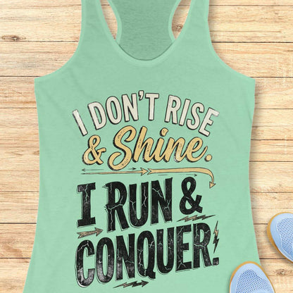 A mint green racerback tank top laid flat on a light wood background, featuring a distressed graphic that reads 'I DON'T RISE & Shine. I RUN & CONQUER!' with decorative arrows and lightning bolts.