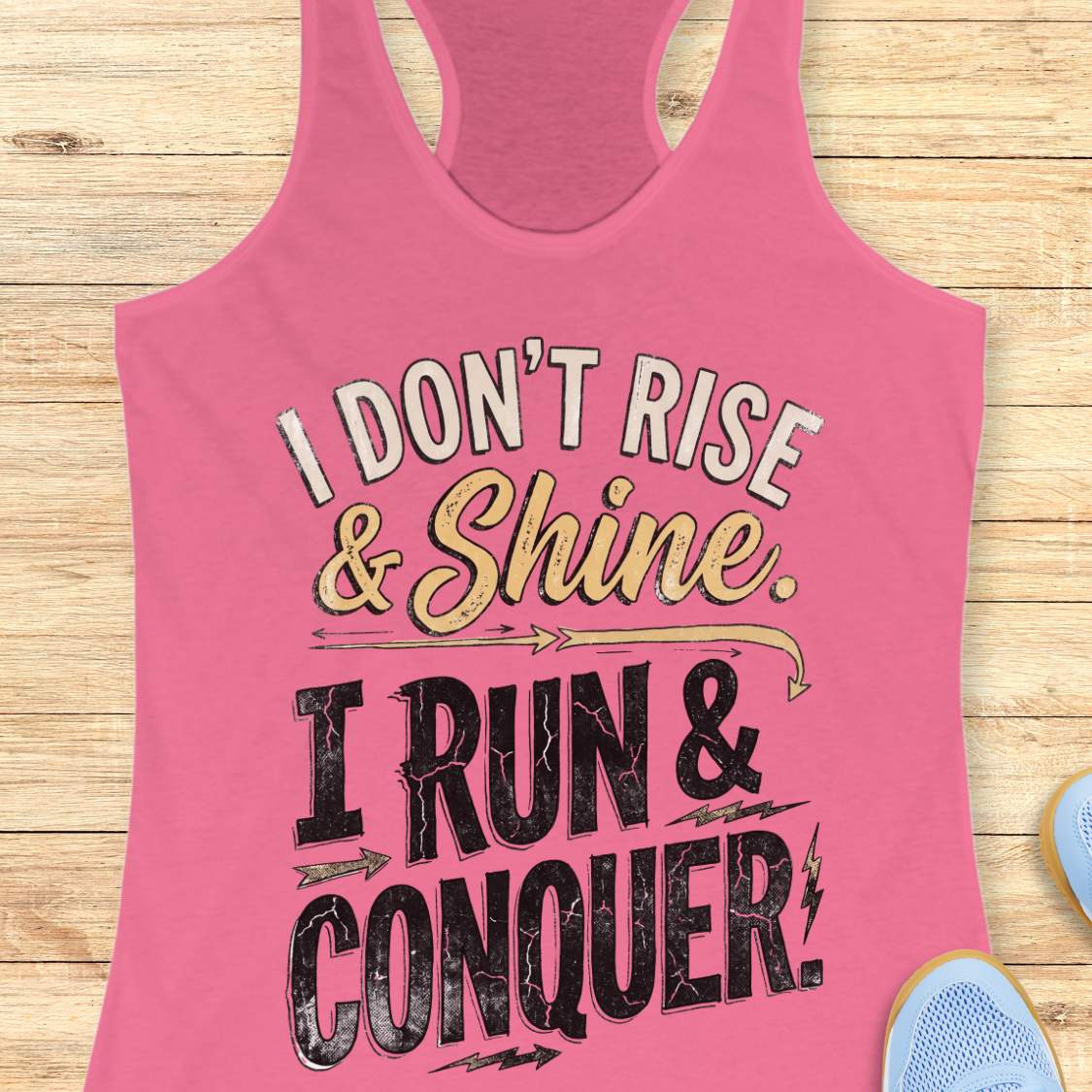 pink racerback running tank top displayed on a light wood background, featuring a distressed vintage-style graphic with blue and white lettering that reads: “I DON’T RISE & SHINE. I RUN & CONQUER.” Decorative arrows and lightning bolt accents surround the text. Partial blue running shoes are visible in the bottom corners of the image.