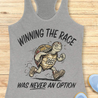 A heather gray racerback tank top laid flat on a light wood surface, featuring a graphic of a turtle runner with the text 'WINNING THE RACE WAS NEVER AN OPTION'.