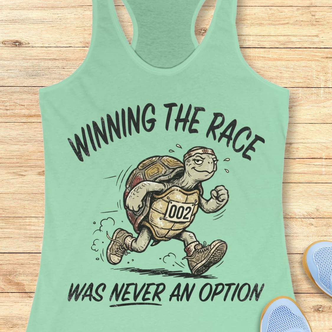mint racerback running tank top displayed on a light wood background, featuring a vintage-style illustration of a turtle running in sneakers and wearing a race bib numbered “002.” The design includes the humorous slogan: “WINNING THE RACE WAS NEVER AN OPTION.” Partial blue running shoes are visible in the bottom corners of the image.