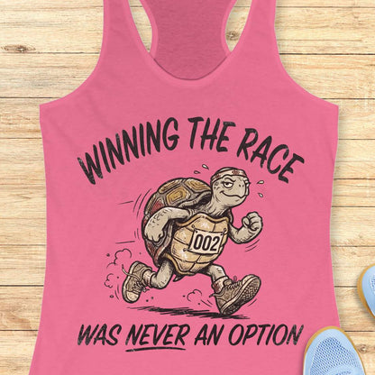 A pink racerback tank top laid flat on a light wood background, featuring a cartoon turtle running with the text 'WINNING THE RACE WAS NEVER AN OPTION'.