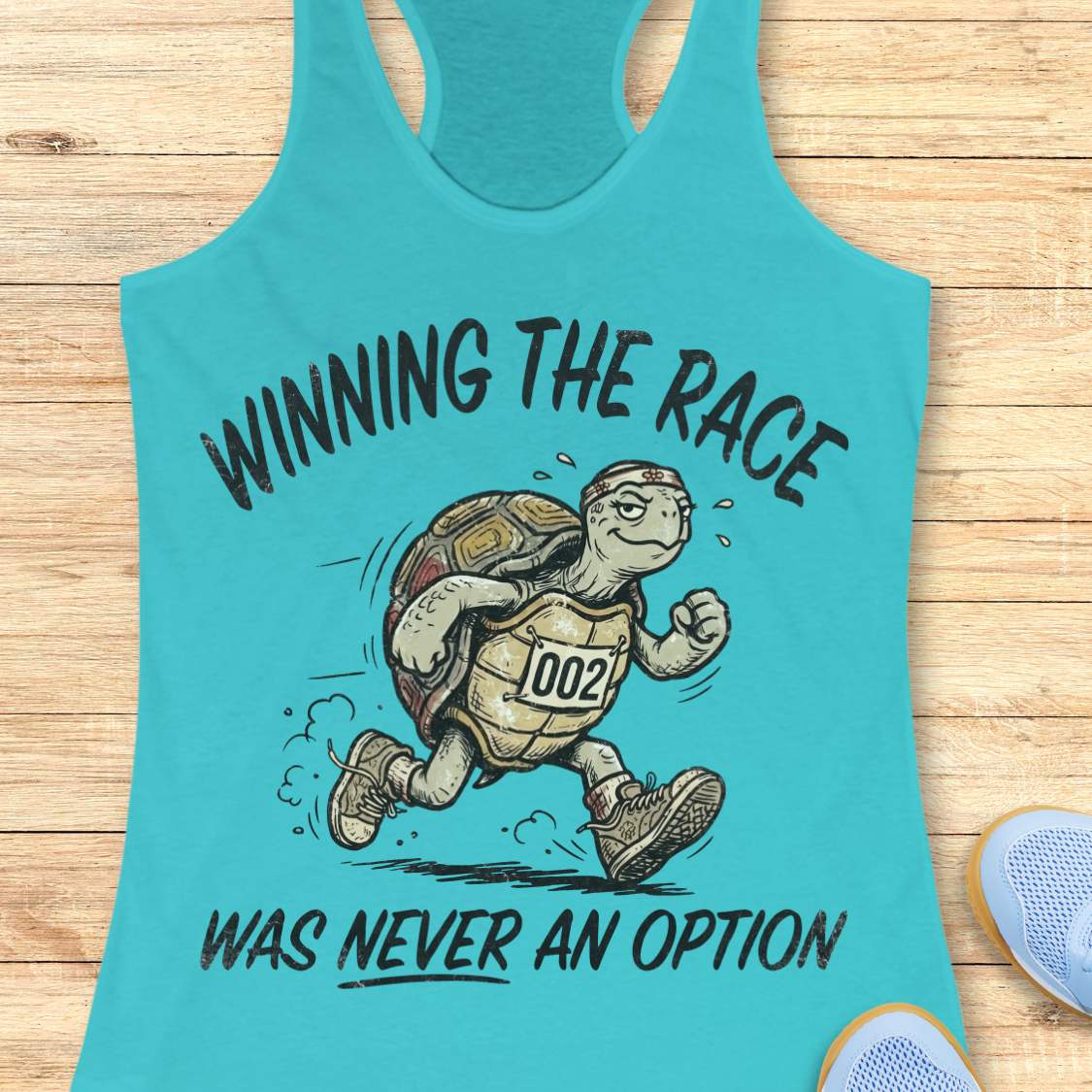 A turquoise racerback tank top laid flat on a light wood surface, featuring a cartoon turtle running with the text 'WINNING THE RACE WAS NEVER AN OPTION.' A partial blue shoe is visible at the lower right edge.