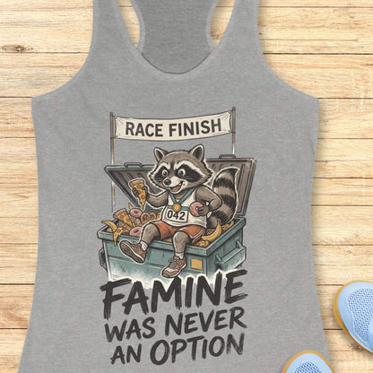 grey racerback running tank top displayed on a light wood background, featuring a vintage-style illustration of a raccoon wearing running shoes, a finisher medal, and race bib number “042.” The raccoon is sitting in an overflowing post-race snack bin filled with pizza, donuts, and bananas beneath a banner that reads “RACE FINISH.” The design includes the humorous slogan: “FAMINE WAS NEVER AN OPTION.” Partial blue running shoes are visible in the bottom corners of the image.