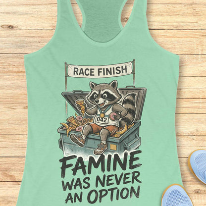 mint racerback running tank top displayed on a light wood background, featuring a vintage-style illustration of a raccoon wearing running shoes, a finisher medal, and race bib number “042.” The raccoon is sitting in an overflowing post-race snack bin filled with pizza, donuts, and bananas beneath a banner that reads “RACE FINISH.” The design includes the humorous slogan: “FAMINE WAS NEVER AN OPTION.” Partial blue running shoes are visible in the bottom corners of the image.
