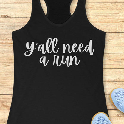 black racerback running tank top displayed on a light wood background, featuring a simple black script design that reads: “Y’all need a run.” The minimalist typography is centered on the front of the tank. Partial blue running shoes are visible in the bottom corners of the image.