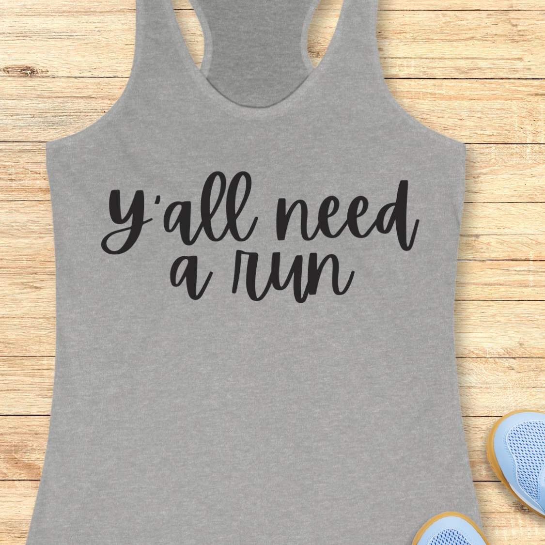 grey racerback running tank top displayed on a light wood background, featuring a simple black script design that reads: “Y’all need a run.” The minimalist typography is centered on the front of the tank. Partial blue running shoes are visible in the bottom corners of the image.