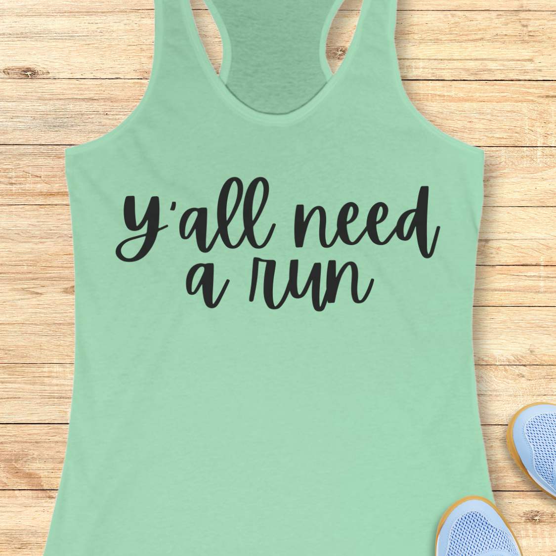 mint racerback running tank top displayed on a light wood background, featuring a simple black script design that reads: “Y’all need a run.” The minimalist typography is centered on the front of the tank. Partial blue running shoes are visible in the bottom corners of the image.