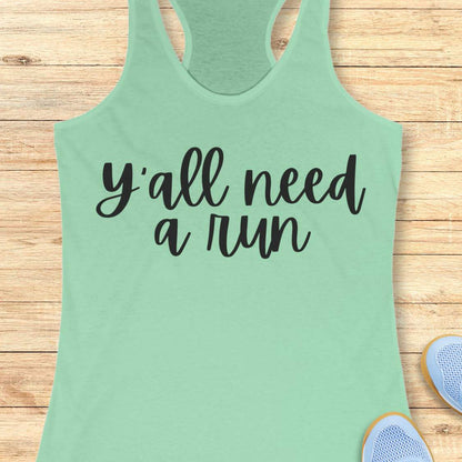 mint racerback running tank top displayed on a light wood background, featuring a simple black script design that reads: “Y’all need a run.” The minimalist typography is centered on the front of the tank. Partial blue running shoes are visible in the bottom corners of the image.