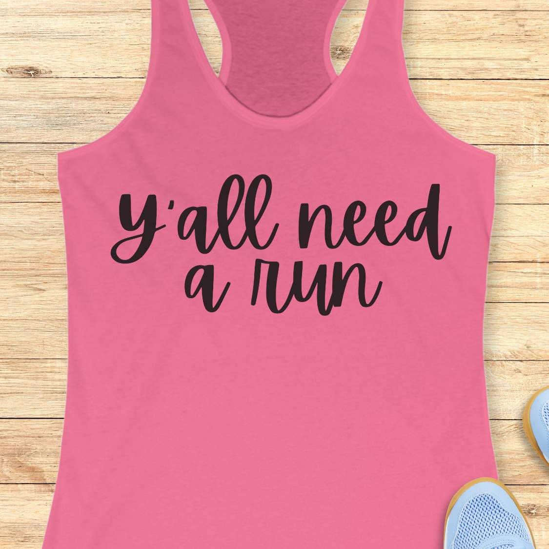 Pink racerback running tank top displayed on a light wood background, featuring a simple black script design that reads: “Y’all need a run.” The minimalist typography is centered on the front of the tank. Partial blue running shoes are visible in the bottom corners of the image.