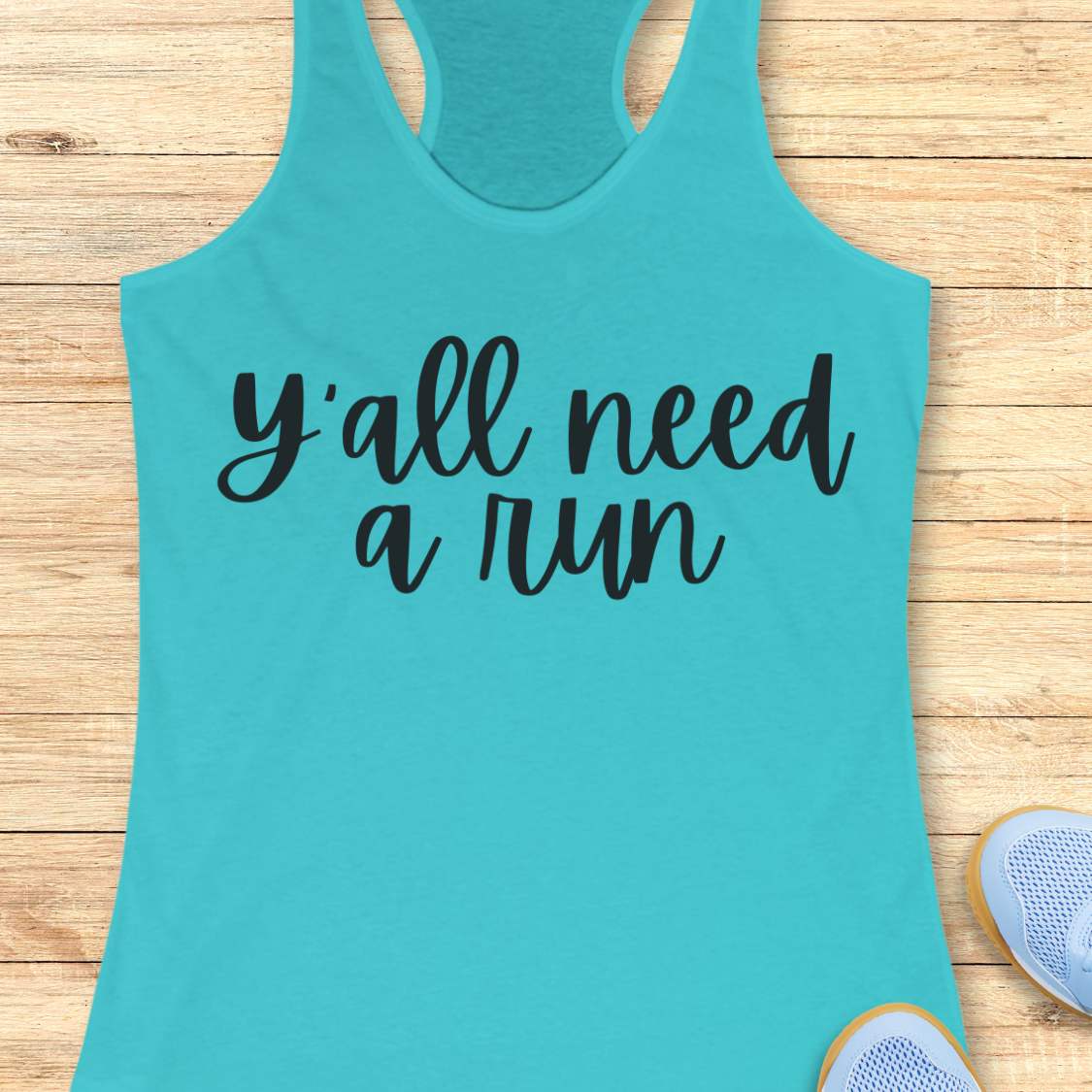 blue racerback running tank top displayed on a light wood background, featuring a simple black script design that reads: “Y’all need a run.” The minimalist typography is centered on the front of the tank. Partial blue running shoes are visible in the bottom corners of the image.