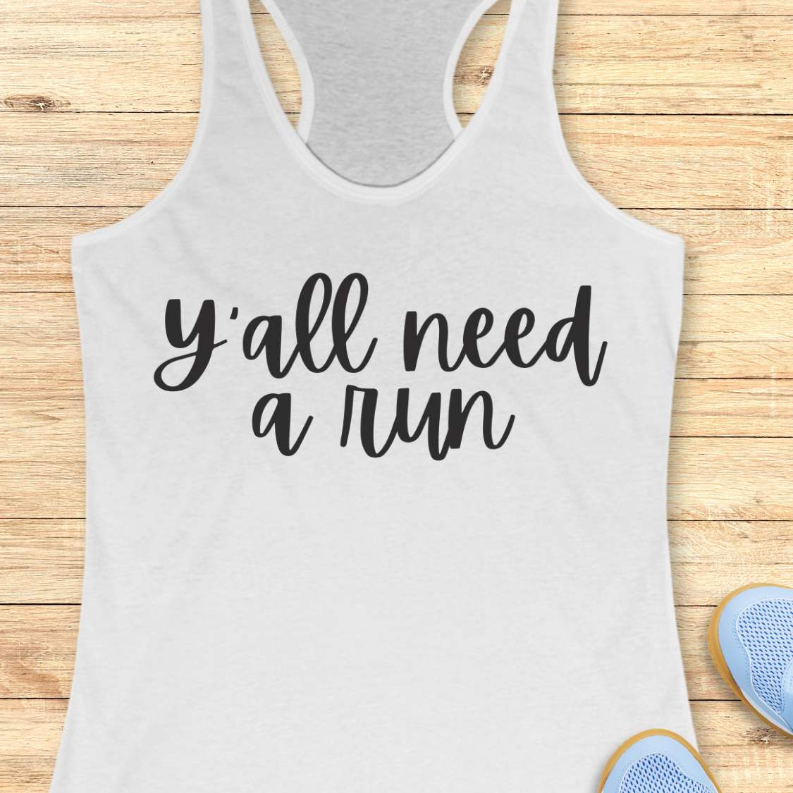 A white racerback tank top with the phrase 'y'all need a run' printed in black script across the front, laid flat on a light wood plank background with partial blue sneakers visible at the bottom right.