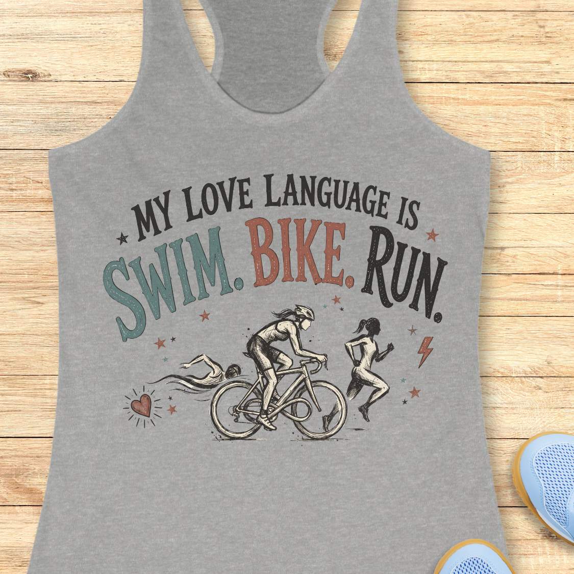 grey racerback tank top displayed on a light wood background, featuring a vintage-inspired triathlon design with illustrations of a swimmer, cyclist, and runner. The graphic includes colorful distressed lettering that reads: “MY LOVE LANGUAGE IS SWIM. BIKE. RUN.” Small decorative stars, a heart, and a lightning bolt accent the design. Partial blue running shoes are visible in the bottom corners of the image.