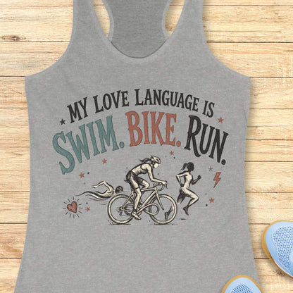 grey racerback tank top displayed on a light wood background, featuring a vintage-inspired triathlon design with illustrations of a swimmer, cyclist, and runner. The graphic includes colorful distressed lettering that reads: “MY LOVE LANGUAGE IS SWIM. BIKE. RUN.” Small decorative stars, a heart, and a lightning bolt accent the design. Partial blue running shoes are visible in the bottom corners of the image.