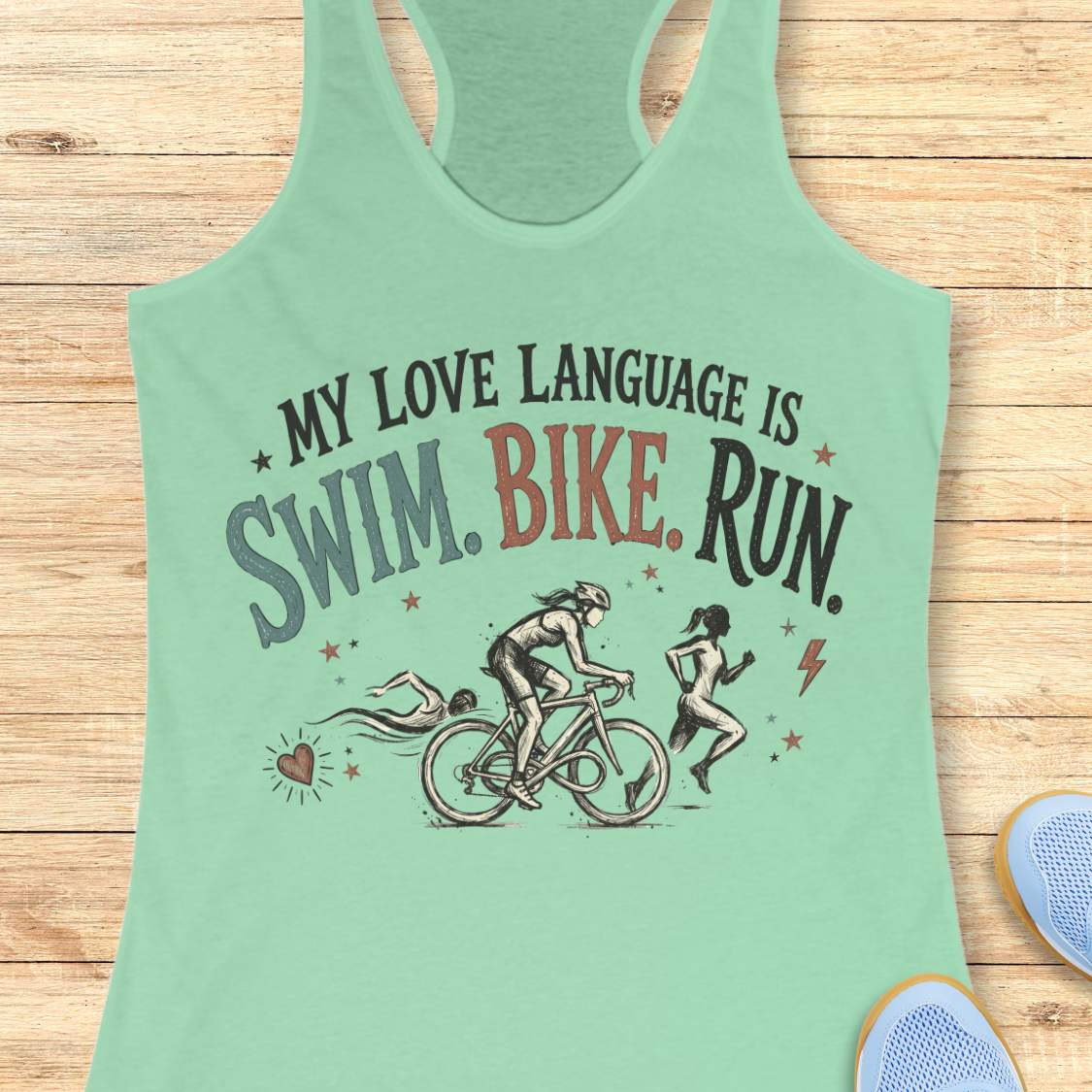 mint racerback tank top displayed on a light wood background, featuring a vintage-inspired triathlon design with illustrations of a swimmer, cyclist, and runner. The graphic includes colorful distressed lettering that reads: “MY LOVE LANGUAGE IS SWIM. BIKE. RUN.” Small decorative stars, a heart, and a lightning bolt accent the design. Partial blue running shoes are visible in the bottom corners of the image.
