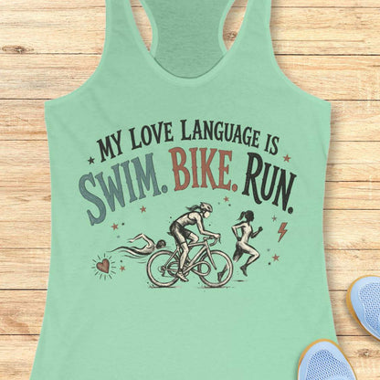 mint racerback tank top displayed on a light wood background, featuring a vintage-inspired triathlon design with illustrations of a swimmer, cyclist, and runner. The graphic includes colorful distressed lettering that reads: “MY LOVE LANGUAGE IS SWIM. BIKE. RUN.” Small decorative stars, a heart, and a lightning bolt accent the design. Partial blue running shoes are visible in the bottom corners of the image.