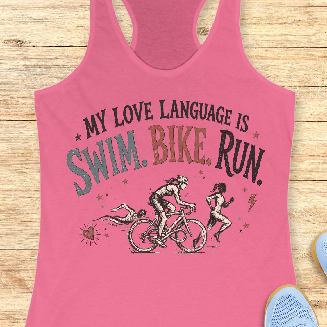 pink racerback tank top displayed on a light wood background, featuring a vintage-inspired triathlon design with illustrations of a swimmer, cyclist, and runner. The graphic includes colorful distressed lettering that reads: “MY LOVE LANGUAGE IS SWIM. BIKE. RUN.” Small decorative stars, a heart, and a lightning bolt accent the design. Partial blue running shoes are visible in the bottom corners of the image.