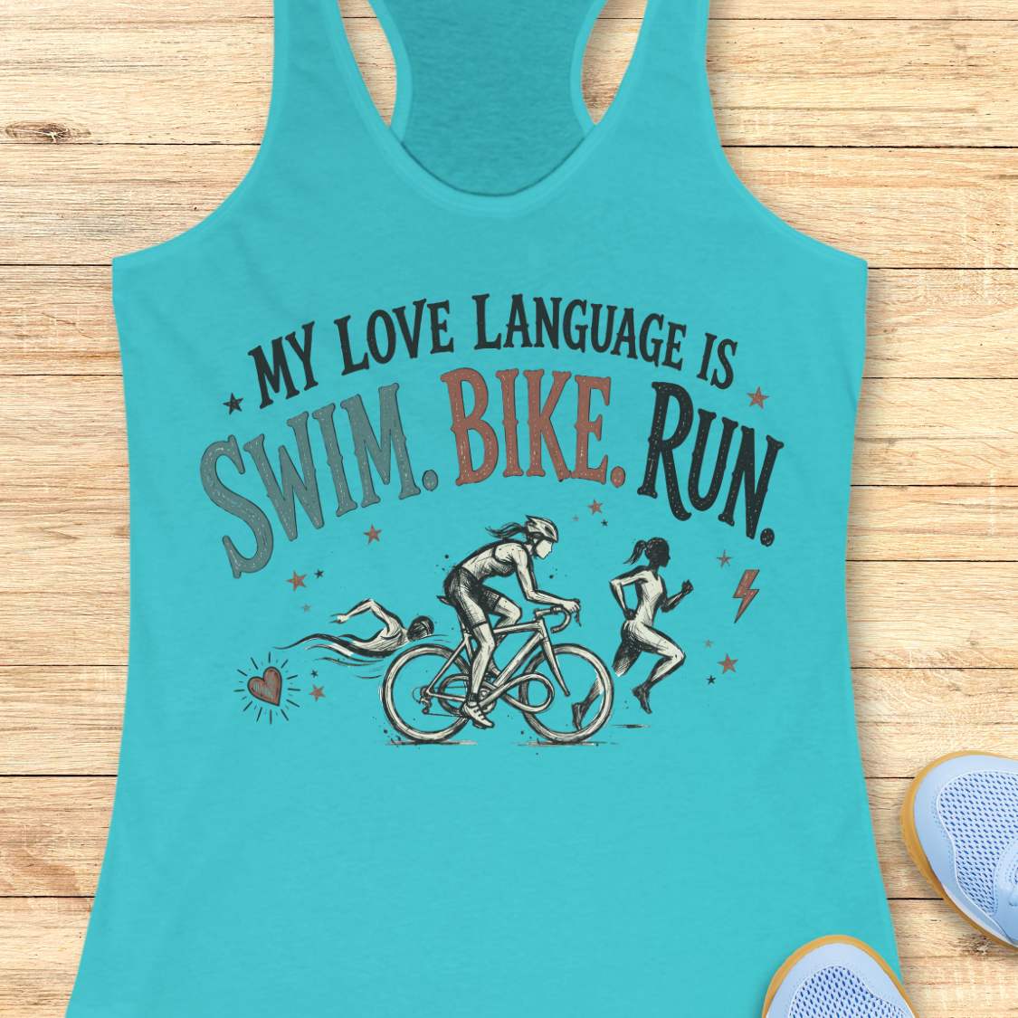 blue racerback tank top displayed on a light wood background, featuring a vintage-inspired triathlon design with illustrations of a swimmer, cyclist, and runner. The graphic includes colorful distressed lettering that reads: “MY LOVE LANGUAGE IS SWIM. BIKE. RUN.” Small decorative stars, a heart, and a lightning bolt accent the design. Partial blue running shoes are visible in the bottom corners of the image.