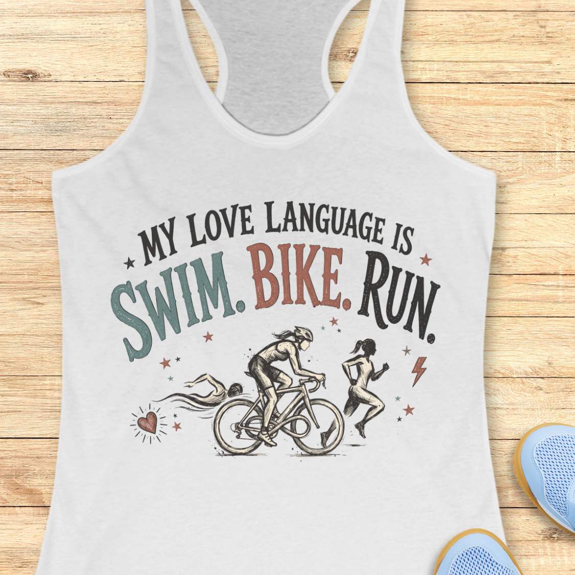 White racerback tank top displayed on a light wood background, featuring a vintage-inspired triathlon design with illustrations of a swimmer, cyclist, and runner. The graphic includes colorful distressed lettering that reads: “MY LOVE LANGUAGE IS SWIM. BIKE. RUN.” Small decorative stars, a heart, and a lightning bolt accent the design. Partial blue running shoes are visible in the bottom corners of the image.