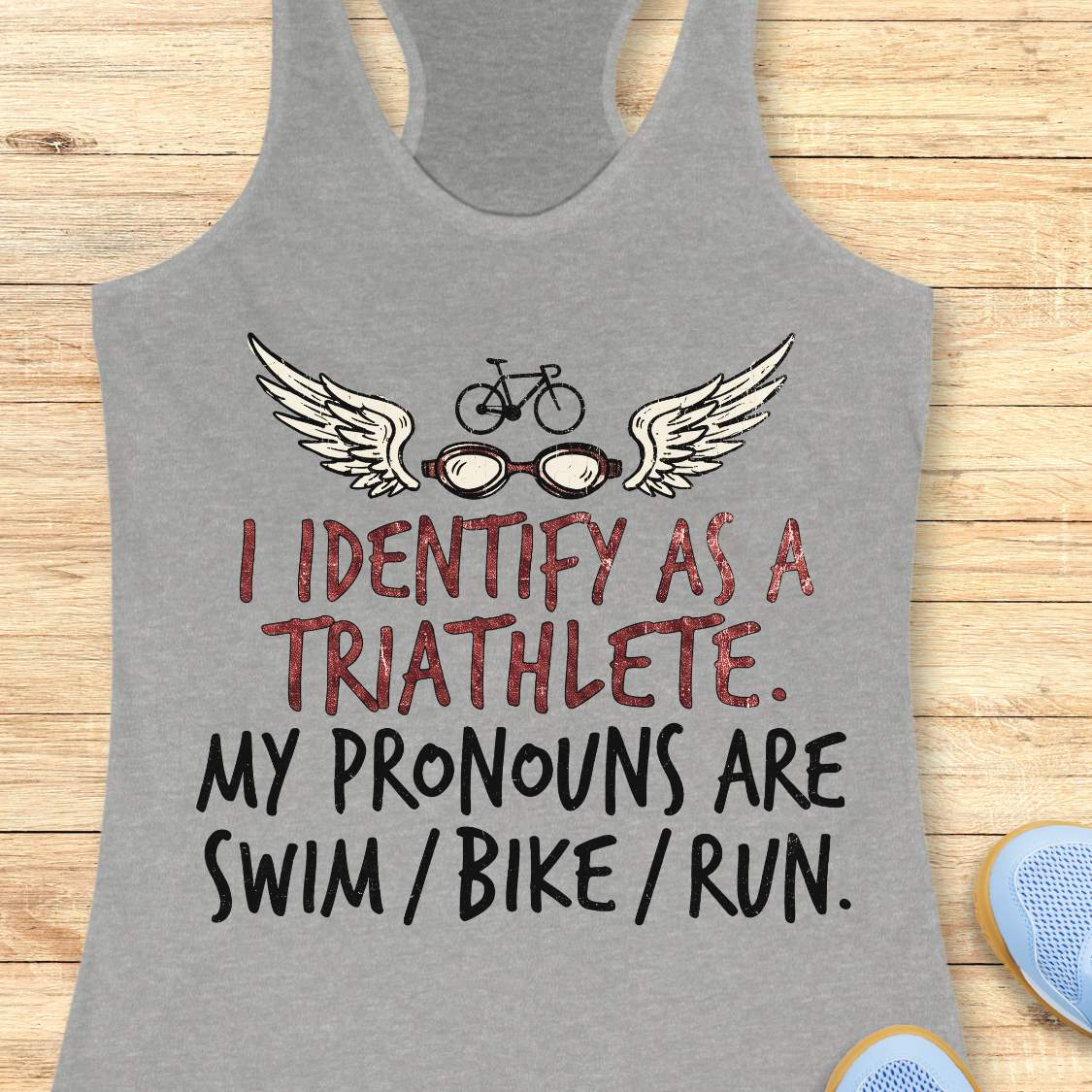 grey racerback tank top displayed on a light wood background, featuring a humorous triathlon-themed design with a bicycle above a pair of winged swim goggles. The graphic includes distressed red and black lettering that reads: “I IDENTIFY AS A TRIATHLETE. MY PRONOUNS ARE SWIM / BIKE / RUN.” Partial blue running shoes are visible in the bottom corners of the image.