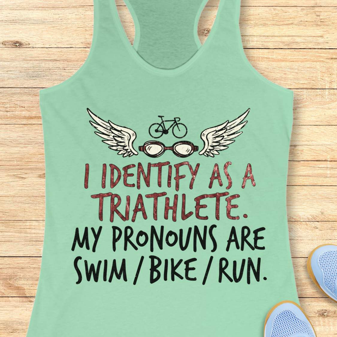 mint racerback tank top displayed on a light wood background, featuring a humorous triathlon-themed design with a bicycle above a pair of winged swim goggles. The graphic includes distressed red and black lettering that reads: “I IDENTIFY AS A TRIATHLETE. MY PRONOUNS ARE SWIM / BIKE / RUN.” Partial blue running shoes are visible in the bottom corners of the image.